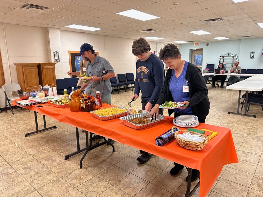 A photo with three people standing in front of an orange fold up table serving themselves food from various dishes on the table.